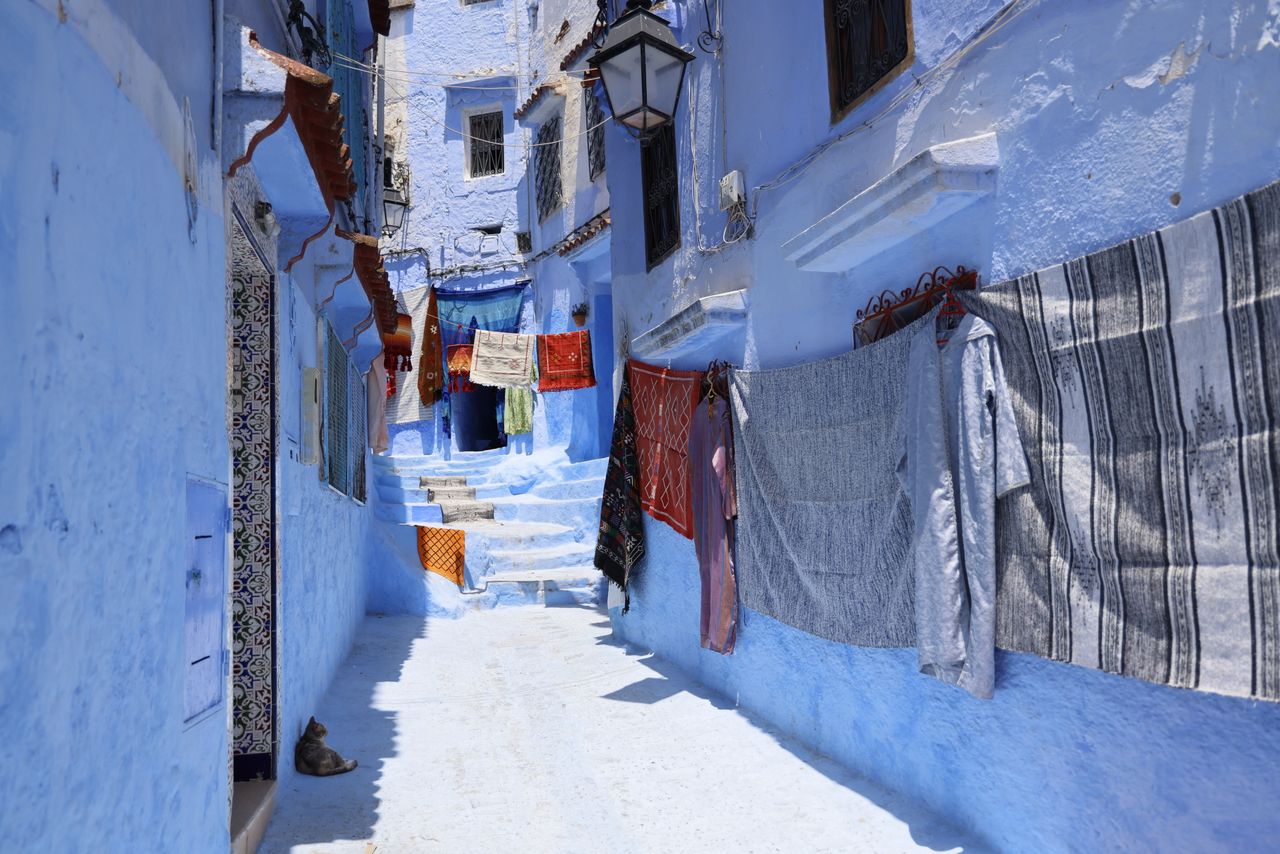 Home Vivid blue alleyway in Chefchaouen, Morocco with colorful drying clothes, vibrant architecture, and sunny ambiance.