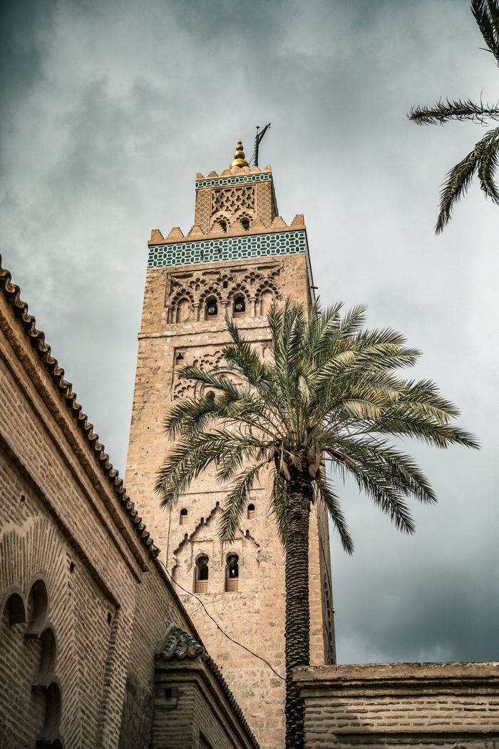Home The iconic Koutoubia Mosque tower with palm trees in Marrakesh, Morocco, under a cloudy sky.